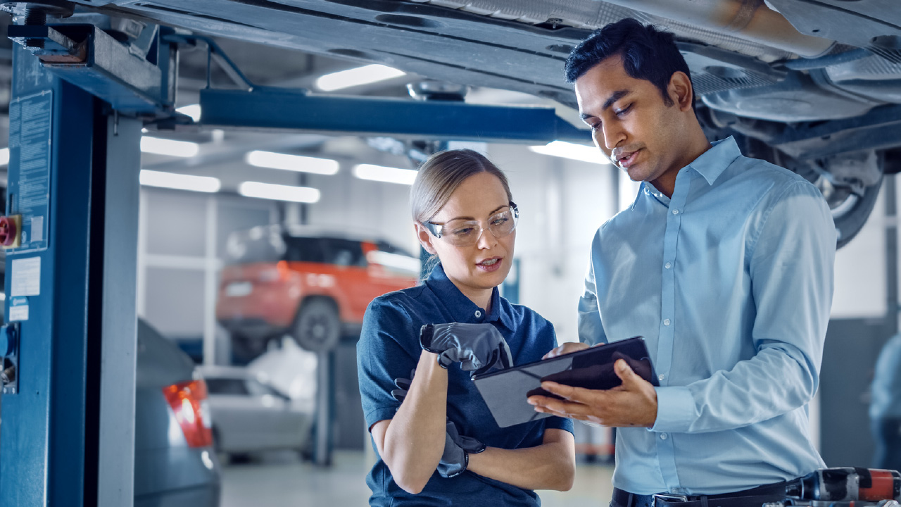  Two mechanics discussing diagnostics under a car lift, demonstrating digital tools for effective automotive asset management.